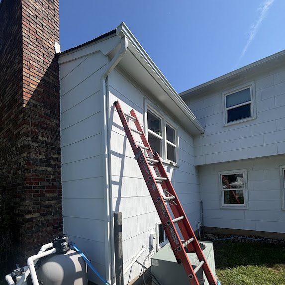 Ladder set at the corner of a white house with a new white downspout beside a brick chimney