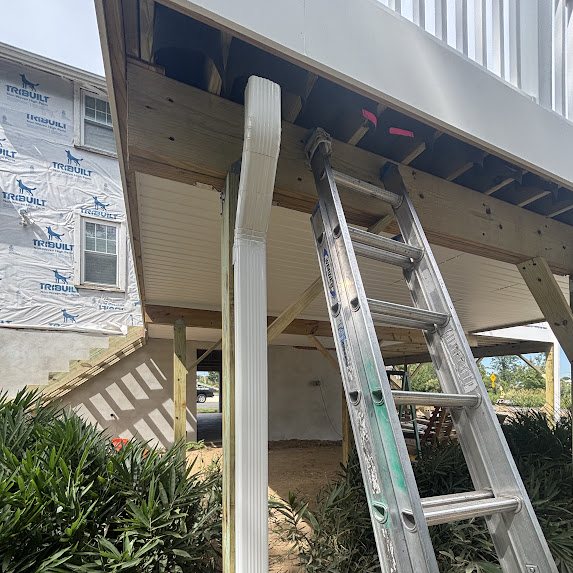 White downspout routed under an elevated deck beside a ladder on a new construction home