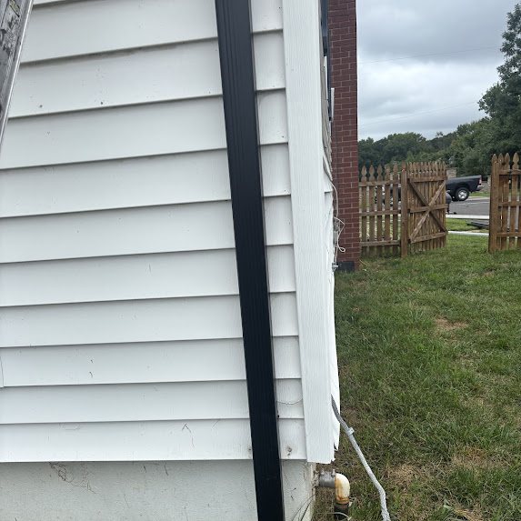 Black downspout running down the corner of a white sided house near a wooden fence