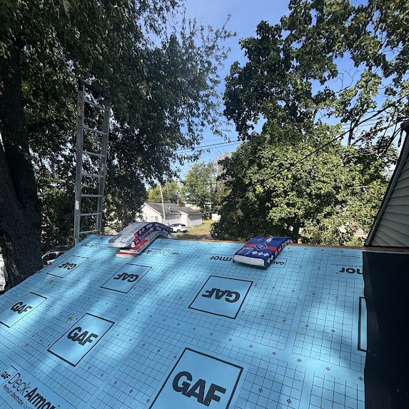 GAF shingle bundles staged on synthetic underlayment beside a ladder